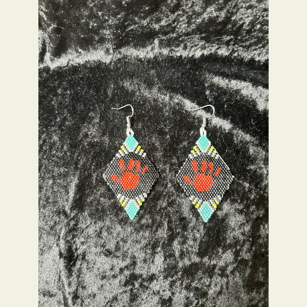 One-of-a-kind MMIW red hand seed bead earrings by Amie Loiselle, featuring a red hand motif on black, turquoise, yellow, and white beaded diamonds, displayed on a black velvet background.