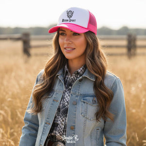 Woman wearing a Bison Spirit limited edition trucker hat, denim jacket, and western shirt standing in a wheat field with a rustic fence in the background