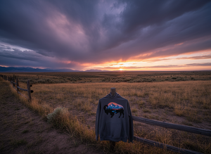 Gray hoodie with colorful bison silhouette and mountain landscape design hanging on a rustic fence at sunset on the Wyoming plains.