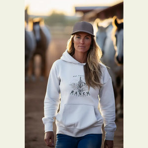 Woman wearing a white Cactus Ranch collection hoodie with horses in the background