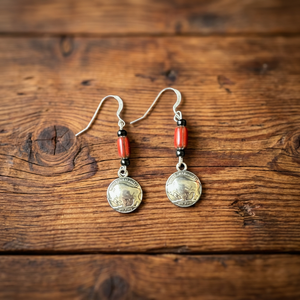 Limited edition earrings by Amie Loiselle featuring red and black trade beads with silver-plated buffalo nickel charms, displayed on rustic wood