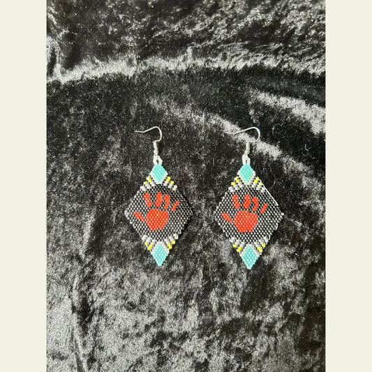 One-of-a-kind MMIW red hand seed bead earrings by Amie Loiselle, featuring a red hand motif on black, turquoise, yellow, and white beaded diamonds, displayed on a black velvet background.