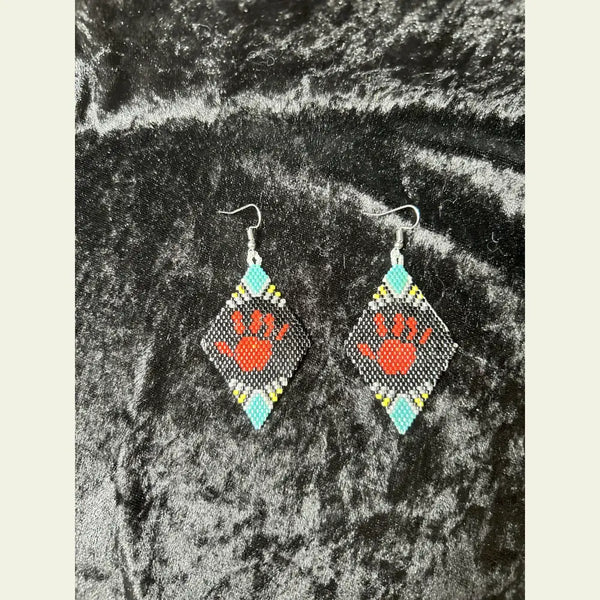 One-of-a-kind MMIW red hand seed bead earrings by Amie Loiselle, featuring a red hand motif on black, turquoise, yellow, and white beaded diamonds, displayed on a black velvet background.