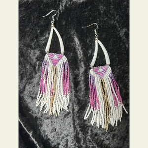 One-of-a-kind long beaded fringe earrings by Amie Loiselle, featuring pink, purple, gold, and white seed beads with shell accents on silver-plated hooks, displayed on a black velvet background.