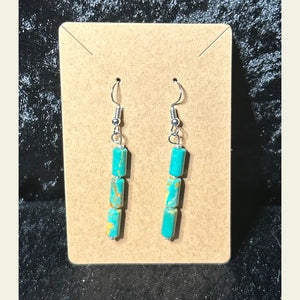 Handcrafted turquoise dangle earrings by artisan Amie Loiselle, featuring three genuine turquoise stones on each earring, displayed on a tan card with a black velvet background.