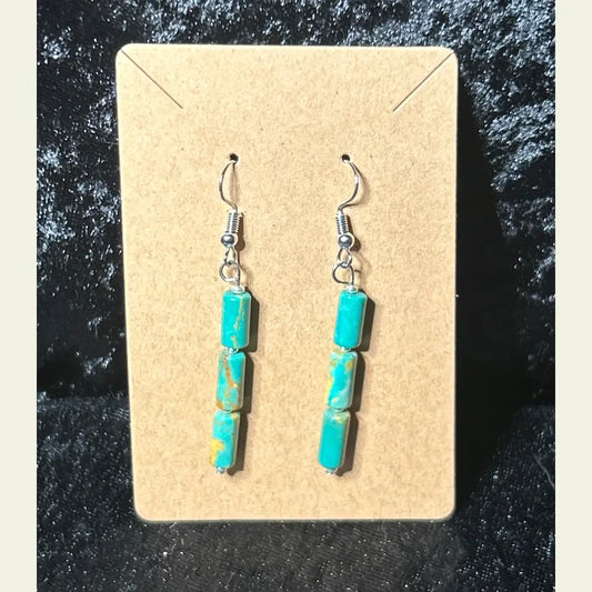 Handcrafted turquoise dangle earrings by artisan Amie Loiselle, featuring three genuine turquoise stones on each earring, displayed on a tan card with a black velvet background.