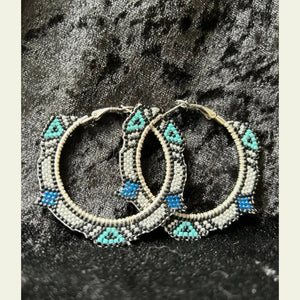 Handcrafted 2-inch seed bead hoop earrings by artisan Amie Loiselle, featuring black, turquoise, white, and blue geometric beadwork, displayed on a black velvet background.