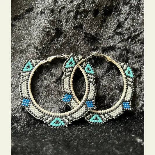 Handcrafted 2-inch seed bead hoop earrings by artisan Amie Loiselle, featuring black, turquoise, white, and blue geometric beadwork, displayed on a black velvet background.